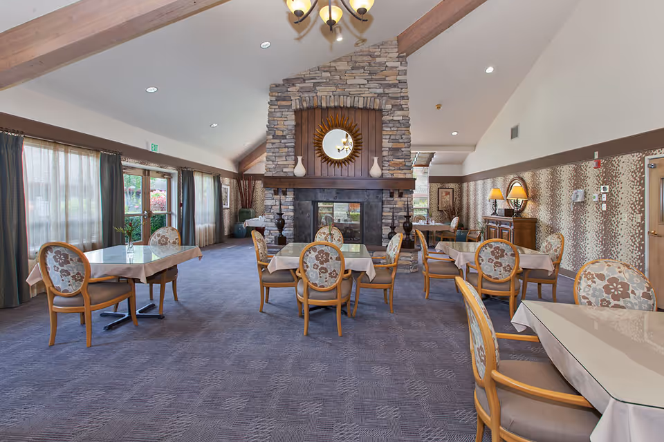 Spacious dining room with table settings and floral-upholstered chairs centered around a large stone fireplace under a vaulted ceiling.