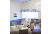 A dining room with several round tables covered with white tablecloths, set with plates, glasses, and napkins. The room has wooden chairs with cushioned seats, a large window with a blue valance, a framed picture on the wall, and a chandelier hanging from the ceiling.
