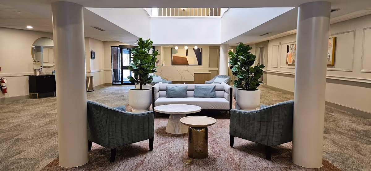 A modern senior living facility lobby with a seating area consisting of a light gray sofa with blue cushions, two blue armchairs, and three small round tables on a patterned rug. Two large potted plants flank the sofa. The space has beige walls with decorative molding, two large white columns, and a reception desk in the background under a skylight.