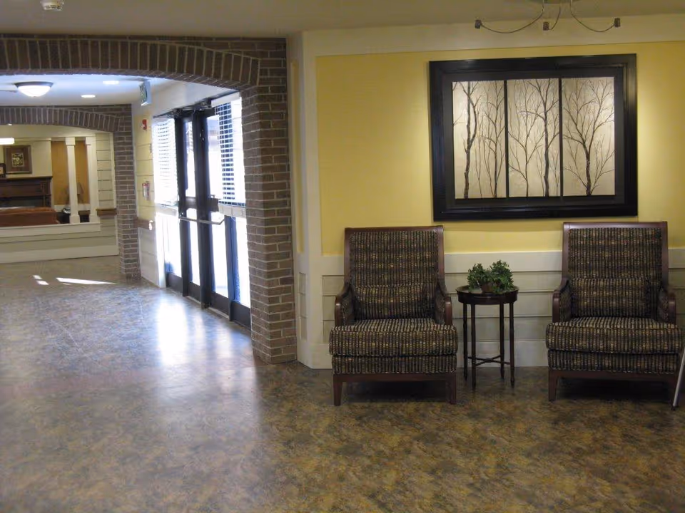 Interior view of a senior living facility hallway with two patterned armchairs separated by a small round table with a plant on it. The walls are painted yellow with white wainscoting, and a framed artwork depicting bare trees hangs above the chairs. To the left, there is a brick archway leading to a set of glass double doors and a glimpse of another room with a fireplace and mirror.