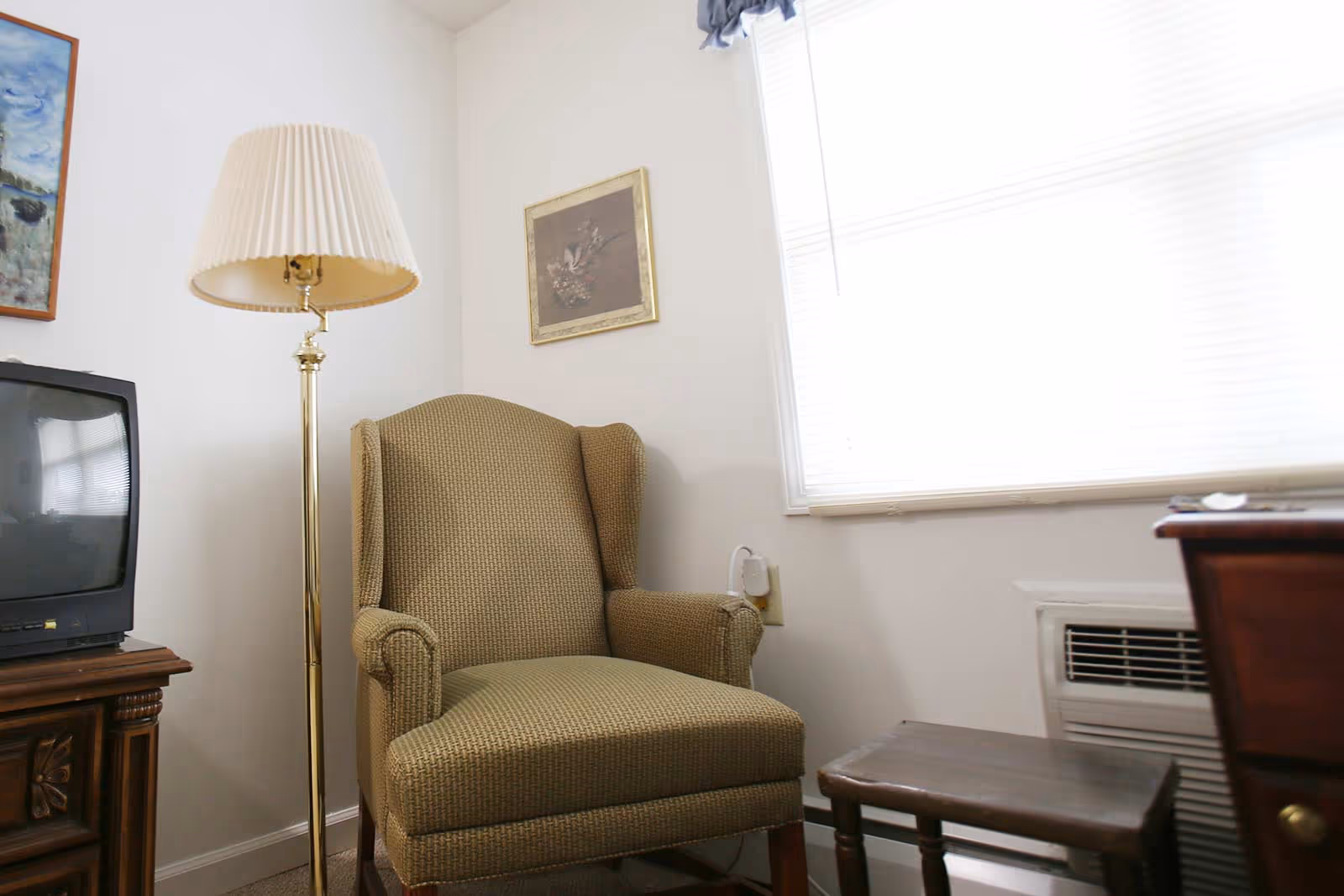 An upholstered wingback chair beside a brass floor lamp, TV and small table next to a bright window.