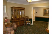 Interior view of a living room area in a senior living facility with patterned carpet, a wooden china cabinet, a striped upholstered chair, a wooden chair, a table lamp, and a painting on the wall.
