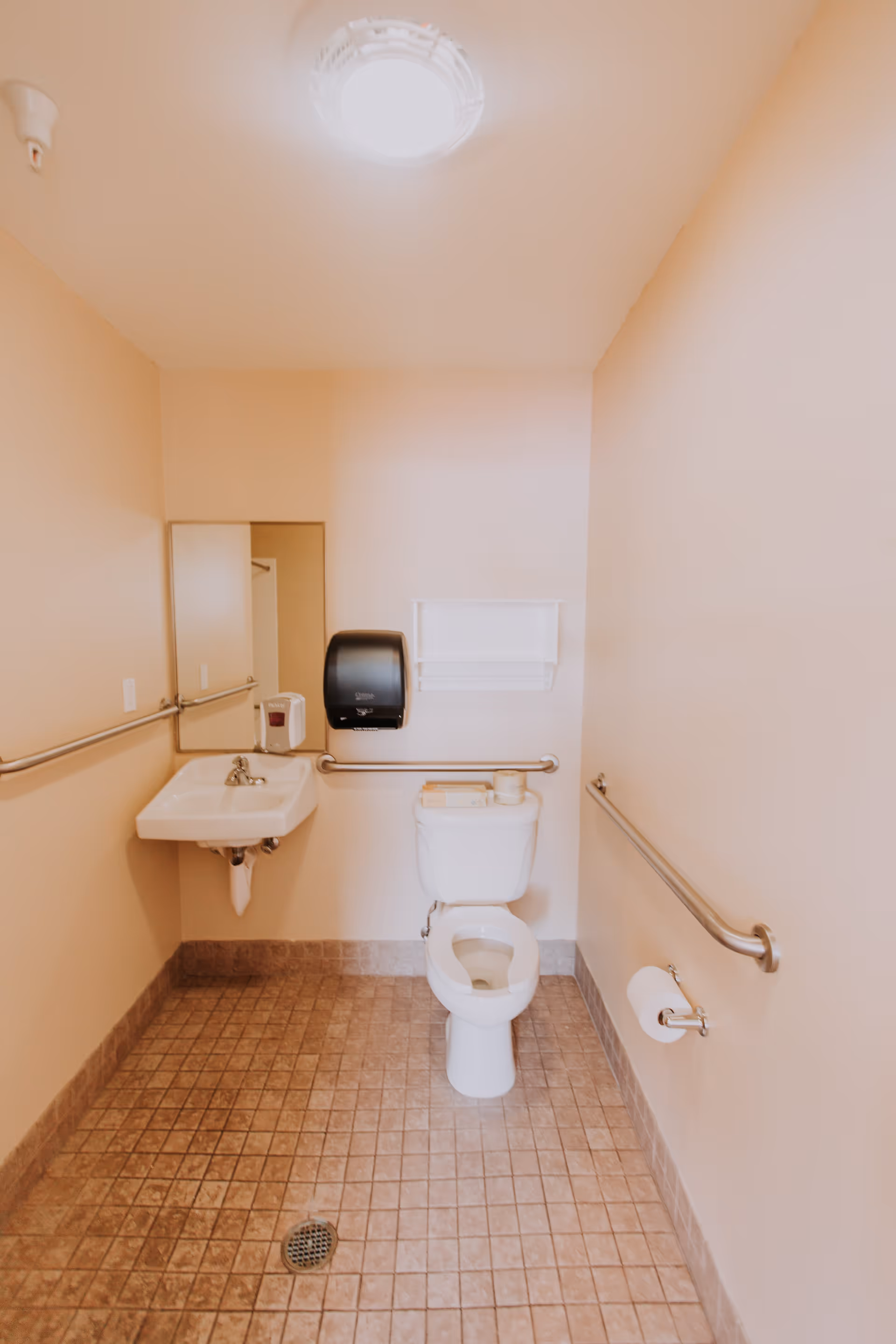 A clean, accessible bathroom with beige walls and tiled floor. The room features a white toilet with the lid open, a small white wall-mounted sink with a faucet, a rectangular mirror above the sink, and stainless steel grab bars on three walls. There is a black paper towel dispenser and a white soap dispenser mounted on the wall above the sink. A toilet paper holder with a roll of toilet paper is mounted on the right wall.