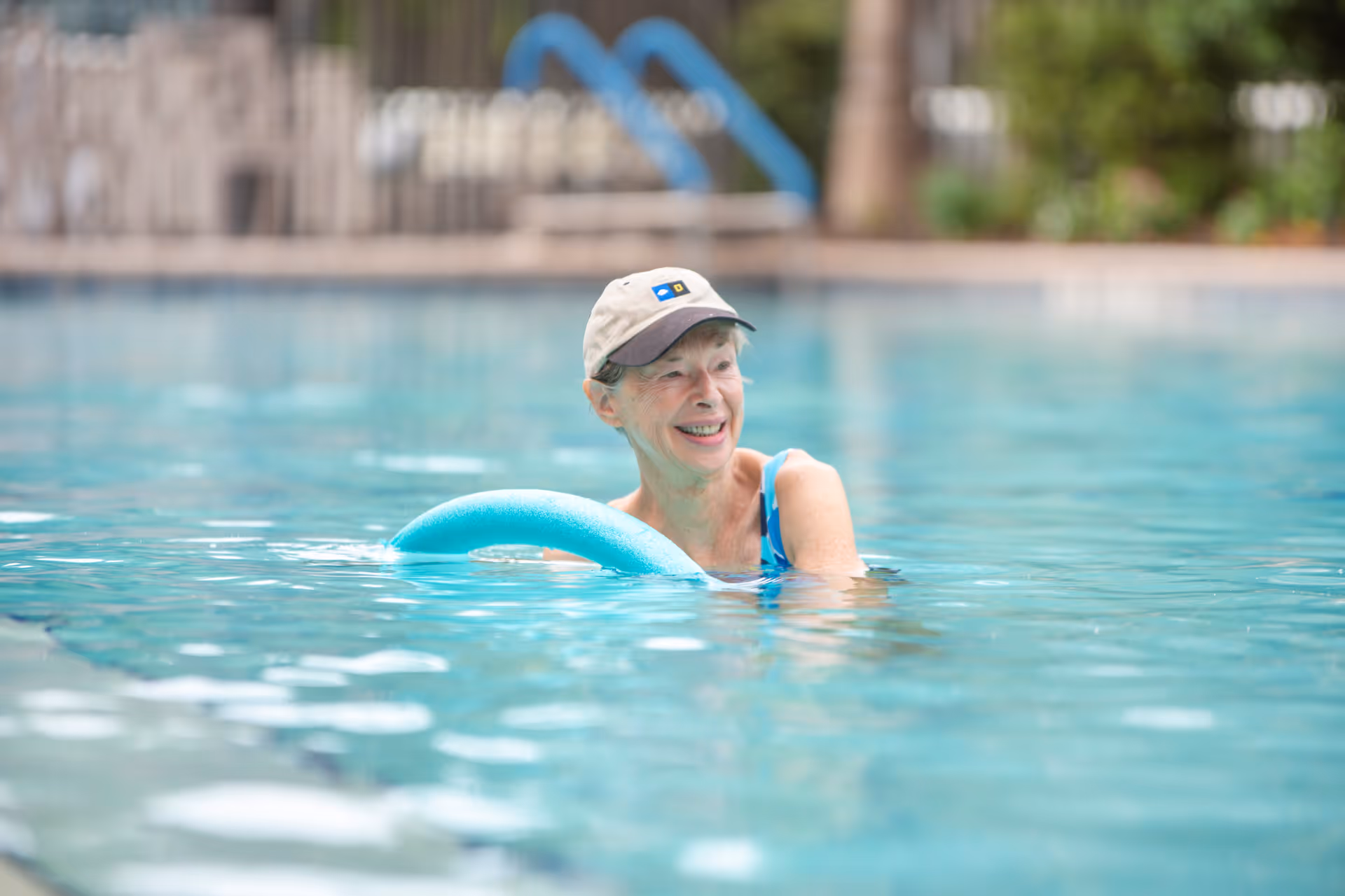 An elderly woman wearing a beige cap and blue swimsuit is smiling while standing in a swimming pool, holding a blue pool noodle. The background shows blurred poolside features including a fence and some greenery.
