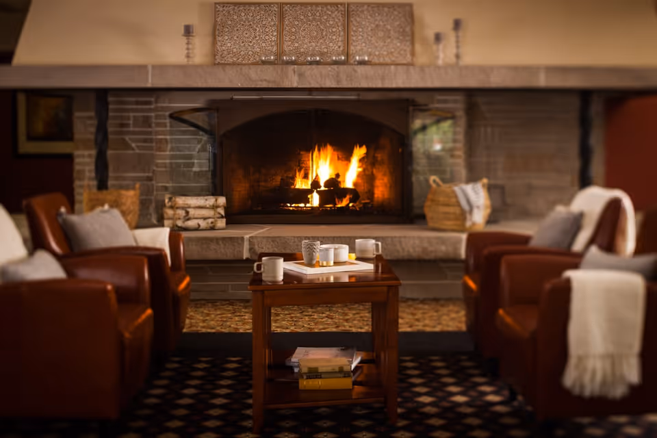 Cozy living room area with a stone fireplace burning a warm fire. Four brown leather armchairs with cushions and throws surround a wooden coffee table that holds mugs and books. Decorative items and baskets are placed on the fireplace mantel and hearth.