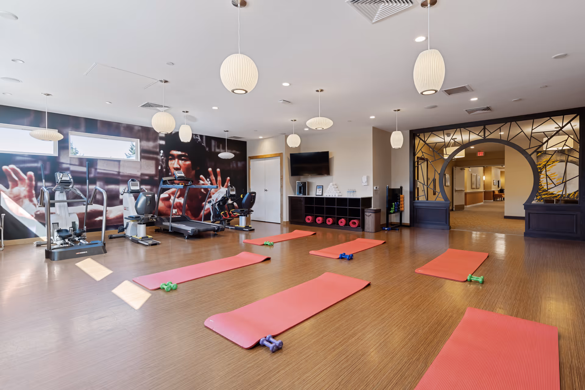 A spacious fitness room with wooden flooring featuring several red exercise mats each paired with small dumbbells. The room has modern exercise equipment including treadmills and elliptical machines along one wall. A large mural of a person in a martial arts pose decorates the back wall. The ceiling has multiple hanging pendant lights, and there is a TV mounted on the wall above a storage unit with pink exercise rollers. An arched decorative doorway leads to a hallway.