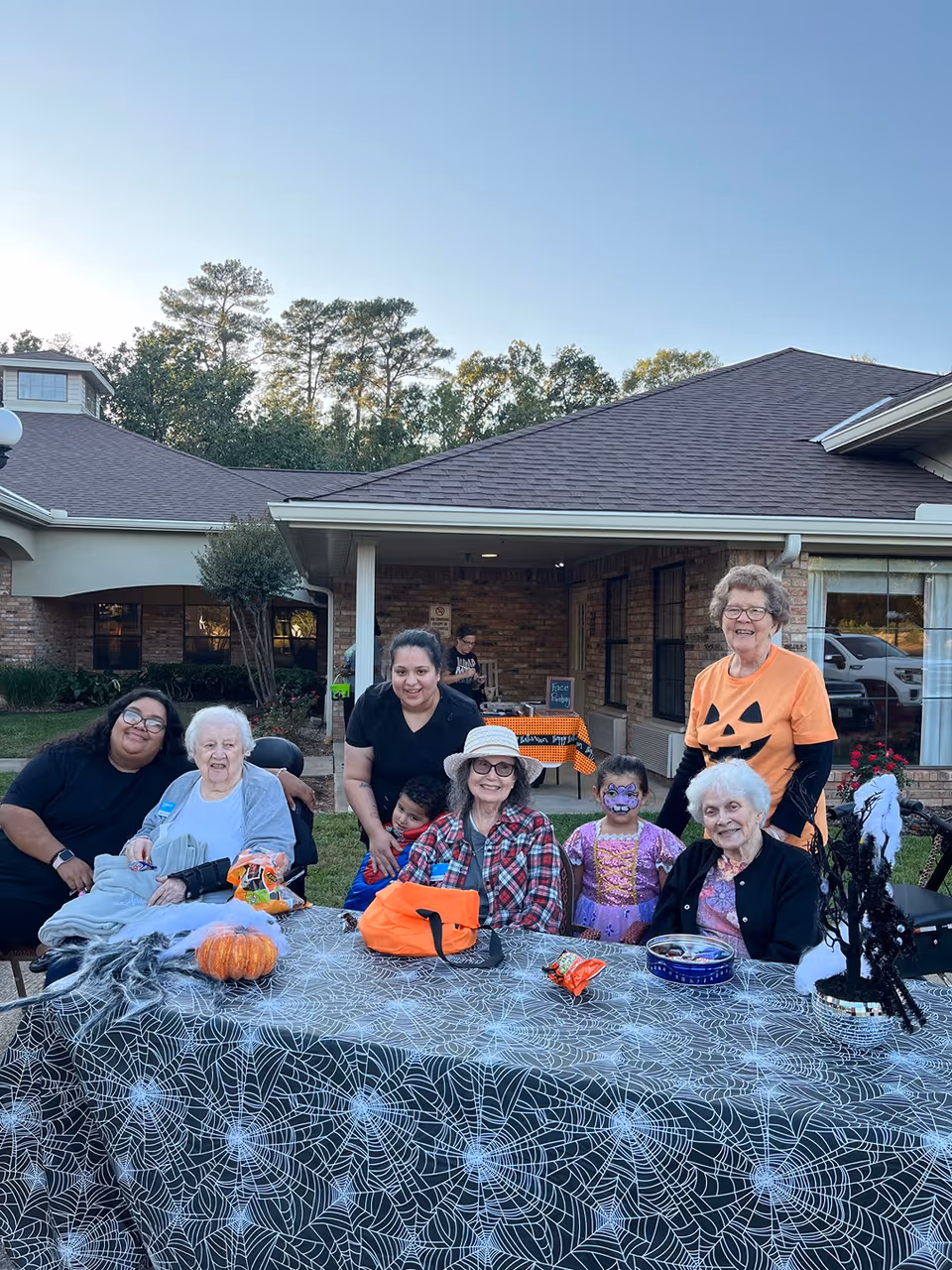 A group of elderly women and two younger adults sitting and standing around a table outdoors decorated with a spider web tablecloth and Halloween-themed items, including a small pumpkin and a black tree with white cobwebs. One child has face paint and is dressed in a purple costume. The setting is in front of a brick building with a covered porch, and the sky is clear.