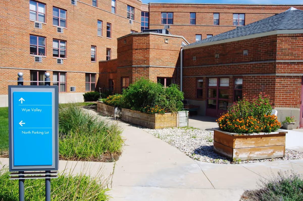 Outdoor courtyard area of a senior living facility with brick buildings surrounding a paved walkway. There are raised garden beds with green plants and orange flowers. A blue directional sign indicates directions to Wye Valley and North Parking Lot.