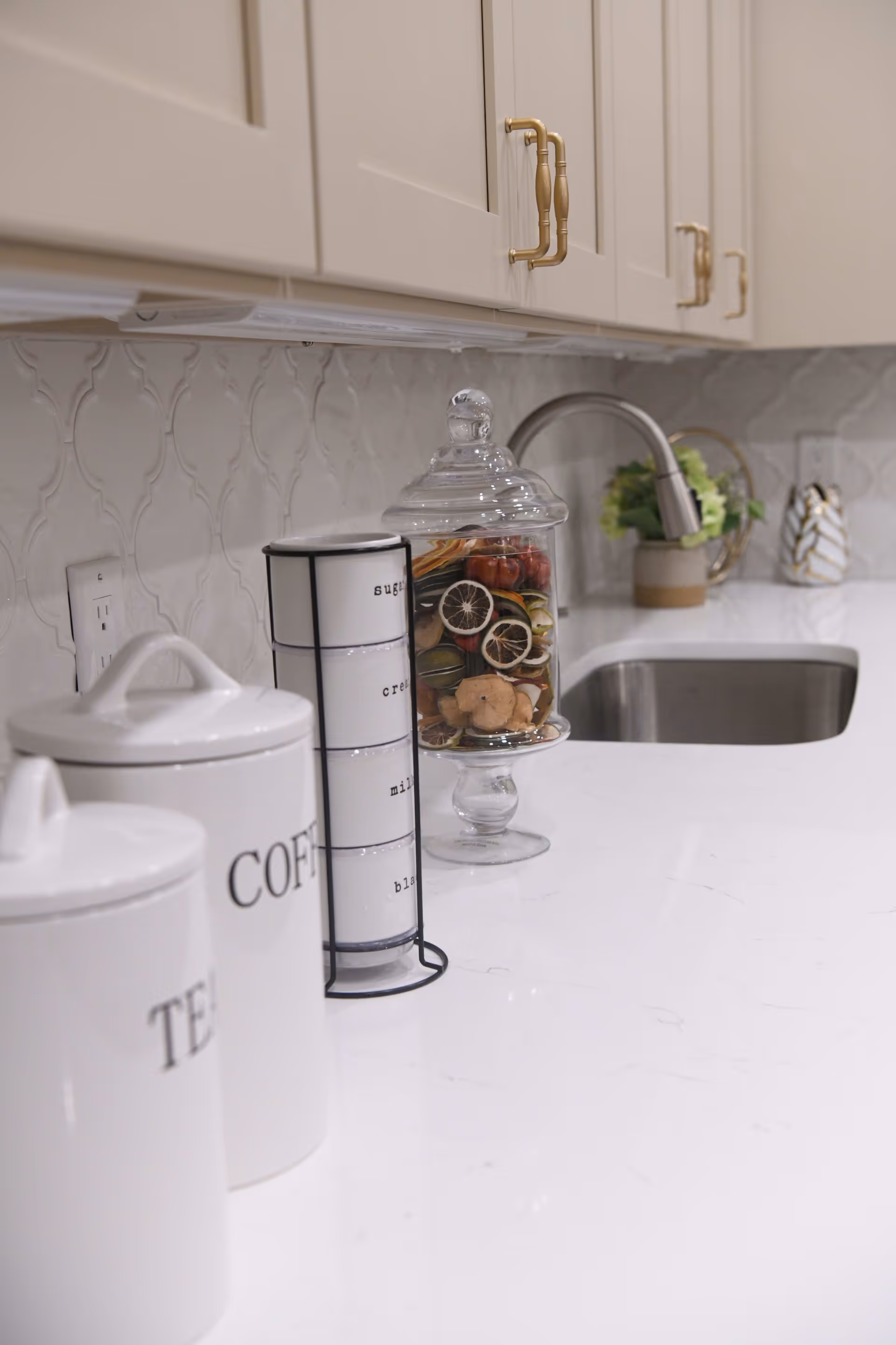 Close-up view of a kitchen countertop with white cabinets above. On the counter are white canisters labeled tea and coffee, a set of stacked white containers labeled sugar, cream, milk, and black, a glass jar filled with dried decorative fruit, and a stainless steel sink with a modern faucet. A small plant and decorative items are placed near the sink.