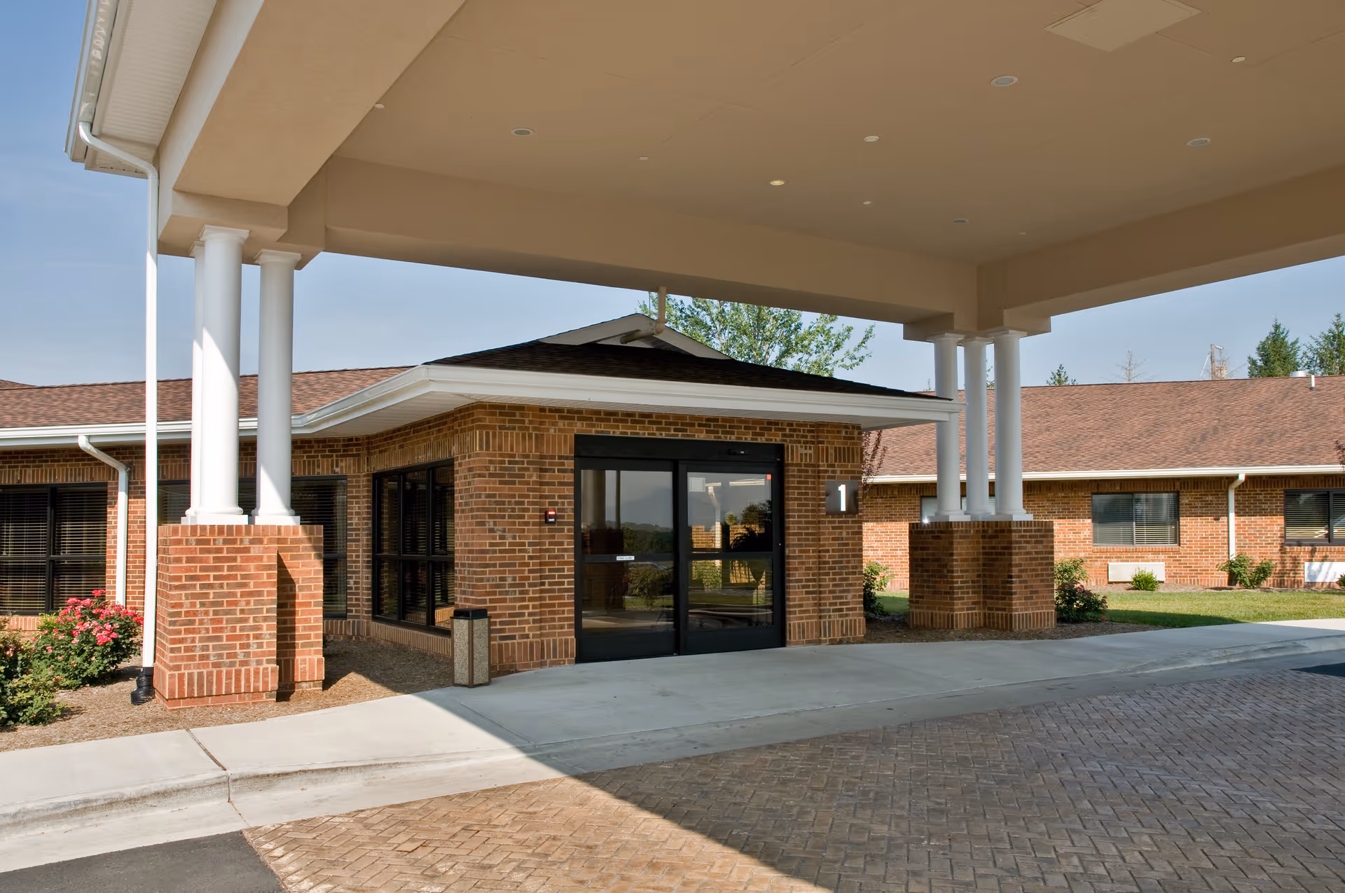Entrance of a brick building with a covered driveway supported by white columns, surrounded by landscaping with bushes and flowers under a clear sky.