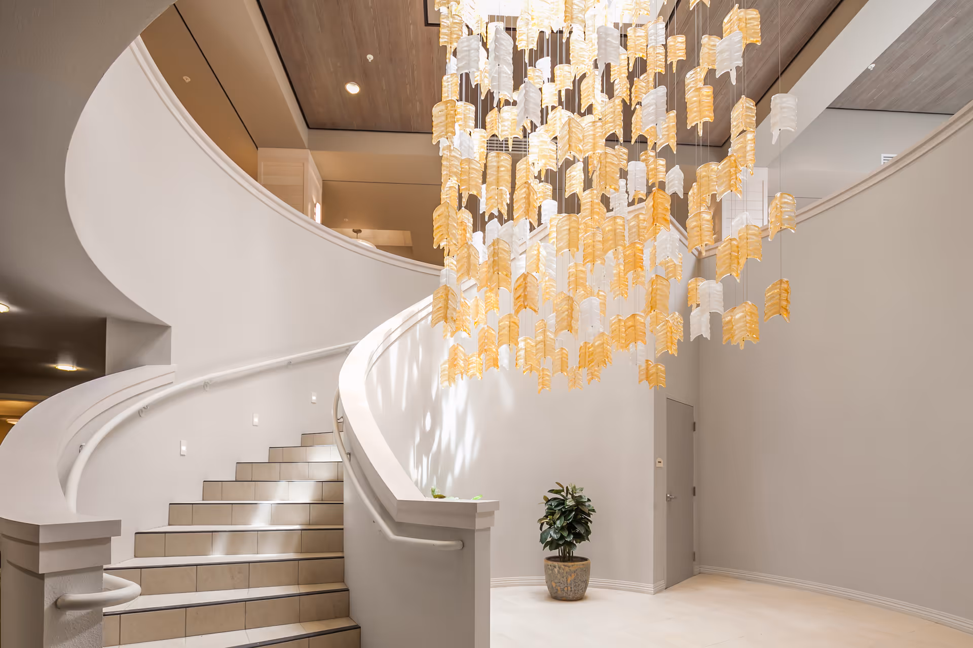 Interior view of a modern senior living facility featuring a curved staircase with white railings and tiled steps. A large decorative chandelier with yellow and white hanging elements is suspended from the ceiling. A potted plant is placed near a closed door on the right side. The walls and floor are light-colored, creating a bright and welcoming atmosphere.