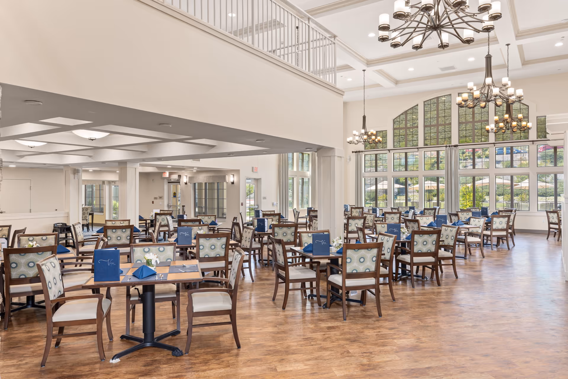 A spacious dining room with multiple wooden tables and chairs arranged neatly. The room features large windows allowing natural light to fill the space, elegant chandeliers hanging from a high ceiling, and menus placed on the tables.