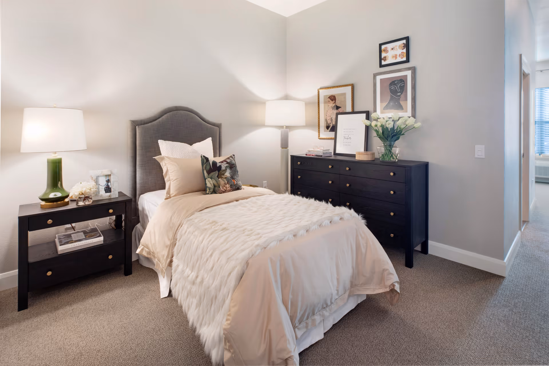 A cozy bedroom with a single bed featuring beige bedding and a fluffy white throw. The bed has a gray upholstered headboard and is flanked by a black nightstand with a green lamp and decorative items. Across from the bed is a black dresser adorned with framed artwork, books, and a vase of white flowers. The room has beige carpet and light-colored walls, with a hallway visible to the right.
