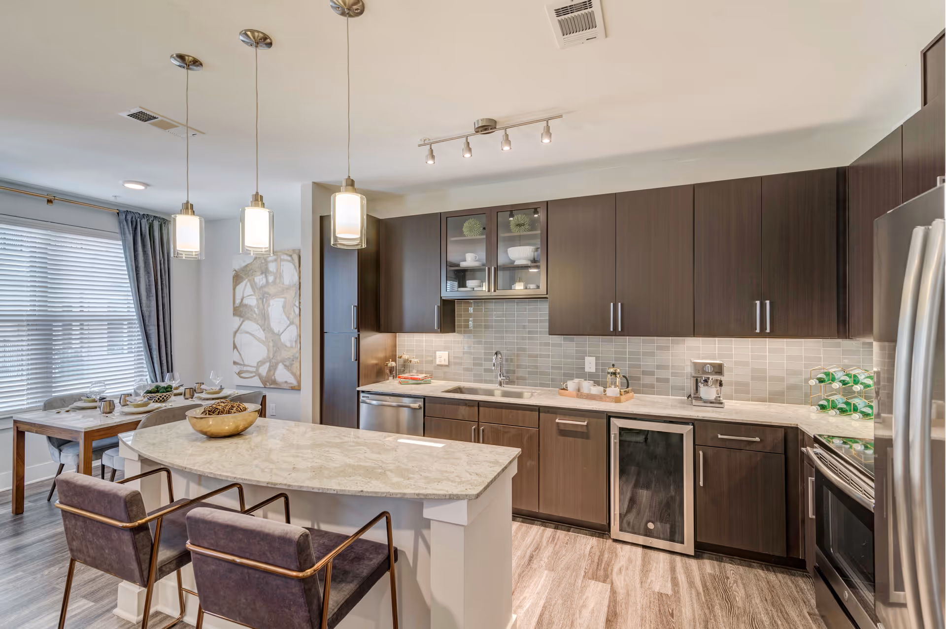 Modern open kitchen with a marble island, dark wood cabinets, pendant lights, and a dining area by a window.