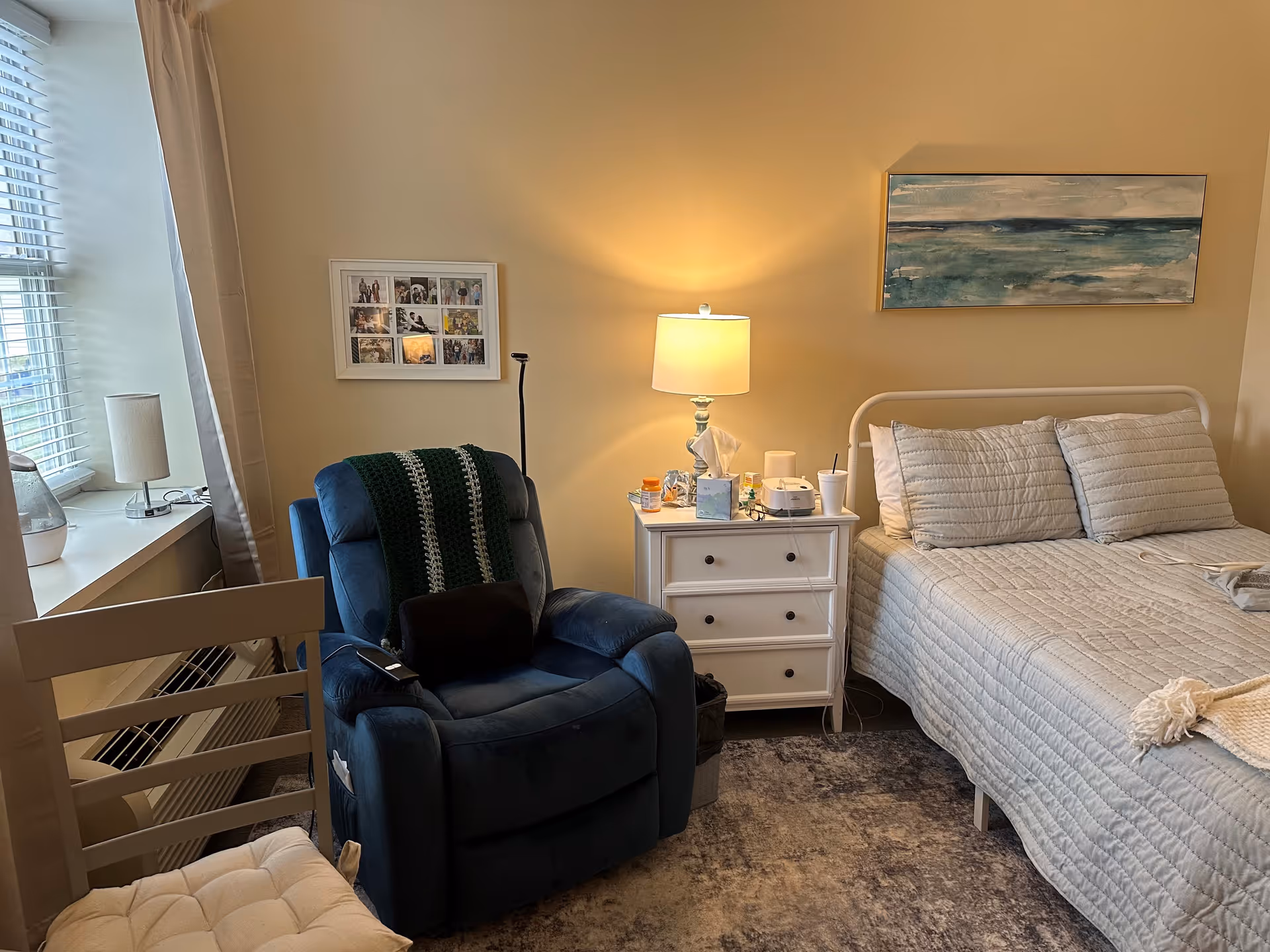 A cozy bedroom with a neatly made bed featuring light gray bedding and two pillows. Next to the bed is a white nightstand with a lamp, tissue box, and various small items. A comfortable blue recliner chair with a green and white crocheted blanket is positioned near the nightstand. A window with blinds and a small lamp on the windowsill is on the left side. The walls are painted beige and decorated with a framed collage of photos and a landscape painting above the bed.