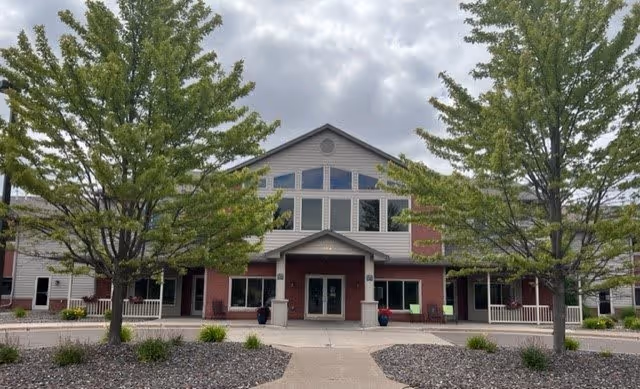 Front exterior view of Duluth Heights Lodge Senior Living building with a pathway leading to the entrance, flanked by two large trees and landscaped areas with rocks and plants under a cloudy sky.