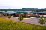 Exterior view of a single-story brick nursing and rehabilitation center building with a green roof, surrounded by a parking lot and some small trees, set against a backdrop of rolling hills and a cloudy sky.