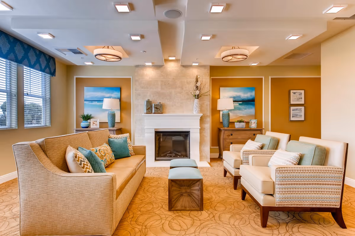 A cozy living room area with a beige sofa adorned with patterned and solid teal cushions, two beige armchairs with light green cushions, and a small ottoman in front of a white fireplace. The walls are painted in warm tones with framed ocean-themed artwork and decorative lamps on wooden side tables. Large windows with blinds allow natural light to fill the room.