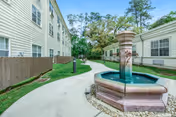 Outdoor walkway between two buildings with a decorative water fountain in the foreground, surrounded by grass and trees in the background under a clear sky.