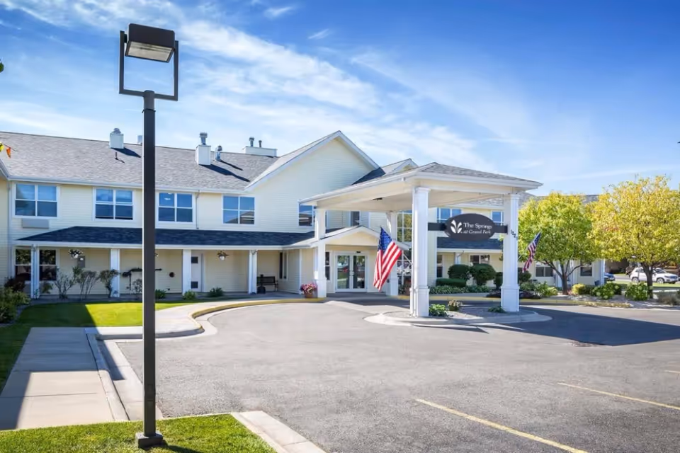 Front entrance of The Springs at Grand Park senior living building with a porte-cochère, American flags, and parking area.