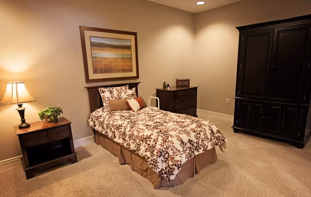 A cozy bedroom with a single bed featuring a floral patterned bedspread and multiple pillows. To the left of the bed is a wooden nightstand with a lamp and a small plant. Above the bed hangs a framed landscape painting. On the right side of the room, there is a dark wooden dresser and a large matching armoire. The room has beige walls and carpeted flooring with recessed ceiling lights.