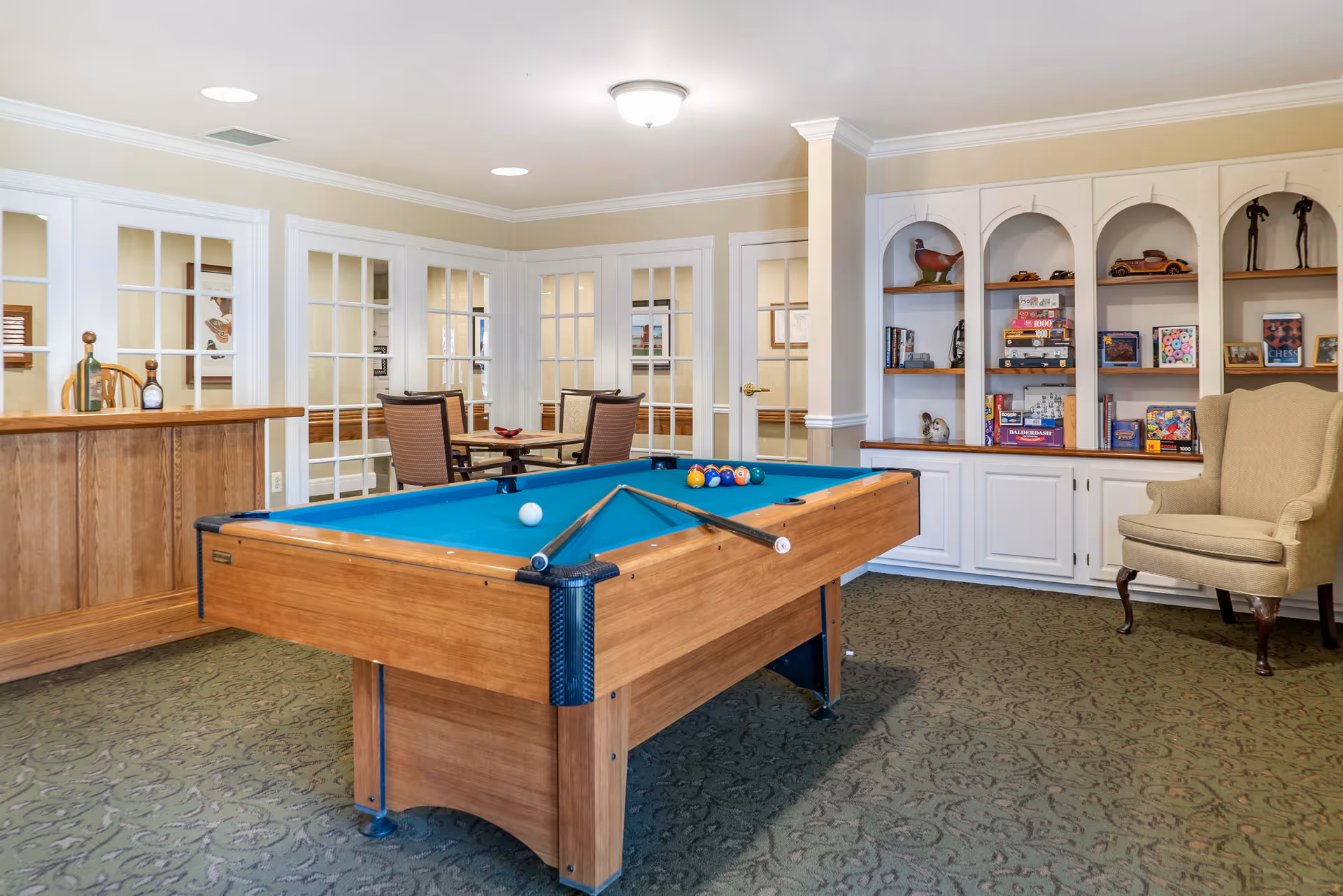 A recreational room with a pool table in the center, two pool cues resting on the table, a small bar area with wooden paneling on the left, a round table with four chairs in the background, and built-in white shelves filled with books, board games, and decorative items on the right. There is also a beige upholstered armchair next to the shelves.