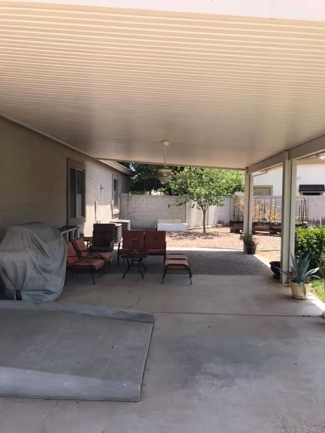 Covered outdoor patio area with seating furniture including chairs and a small table, a covered grill, ceiling fan, and a view of a backyard with a tree, garden beds, and a concrete ramp.