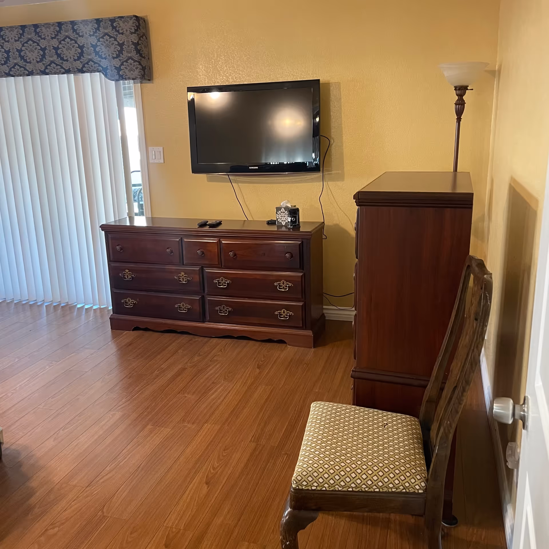 Interior view of a room with wooden flooring, a wall-mounted flat-screen TV above a wooden dresser, a tall wooden cabinet, a cushioned wooden chair, a floor lamp, and vertical blinds covering a sliding glass door.