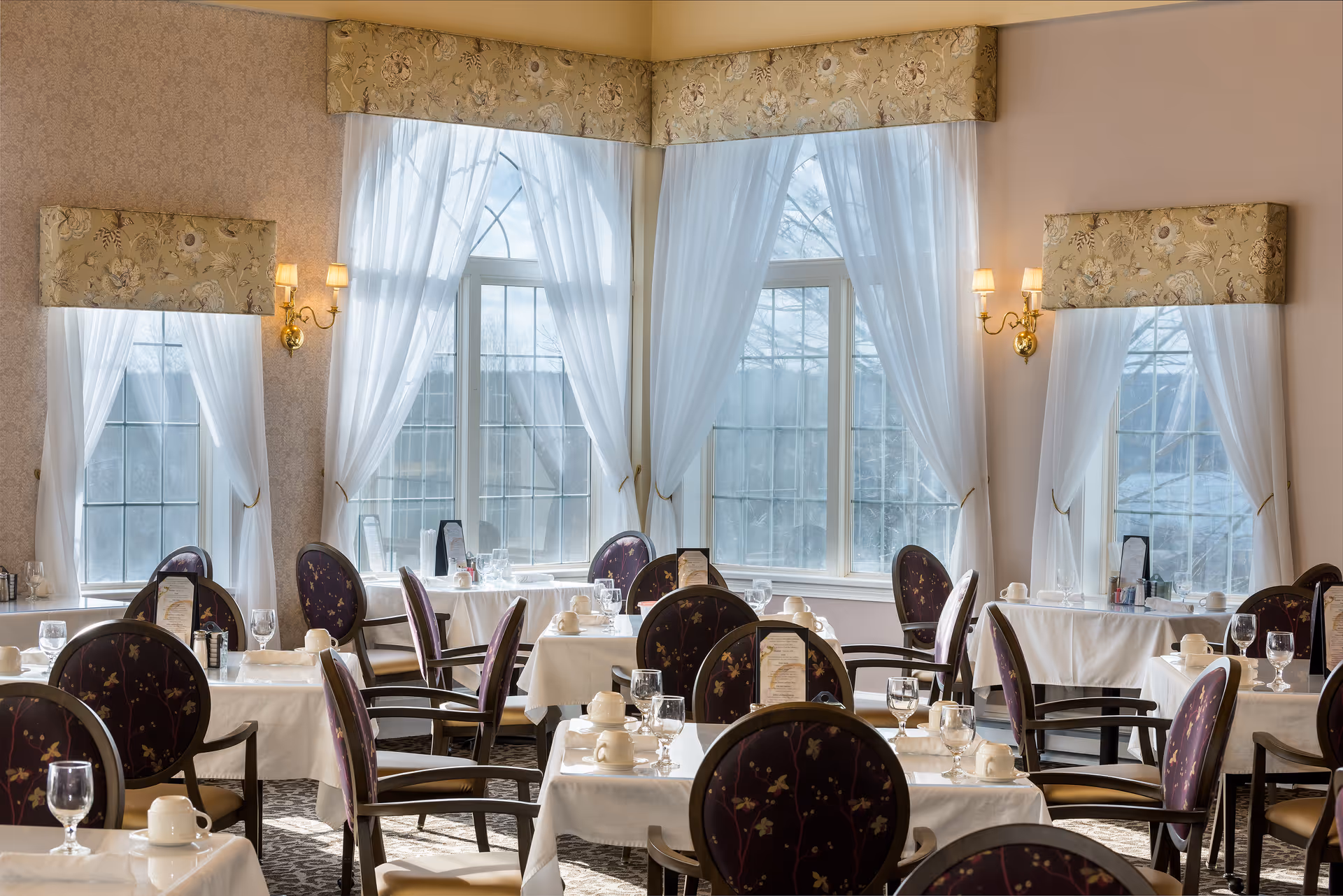 A bright dining room with tables covered in white linens, set with glassware and upholstered chairs in front of tall draped windows.