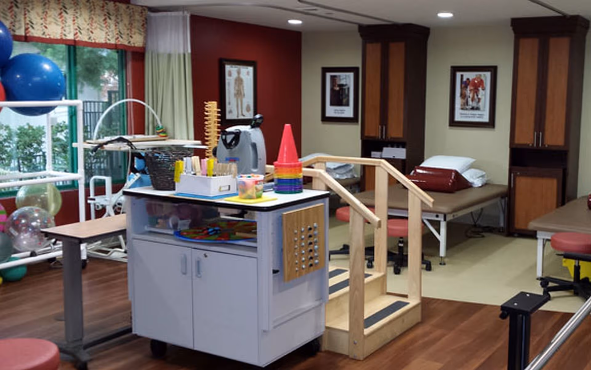 A therapy or rehabilitation room with exercise equipment, therapy tables with pillows, a small wooden staircase with handrails, colorful cones, therapy balls, and framed anatomical and motivational posters on the walls.
