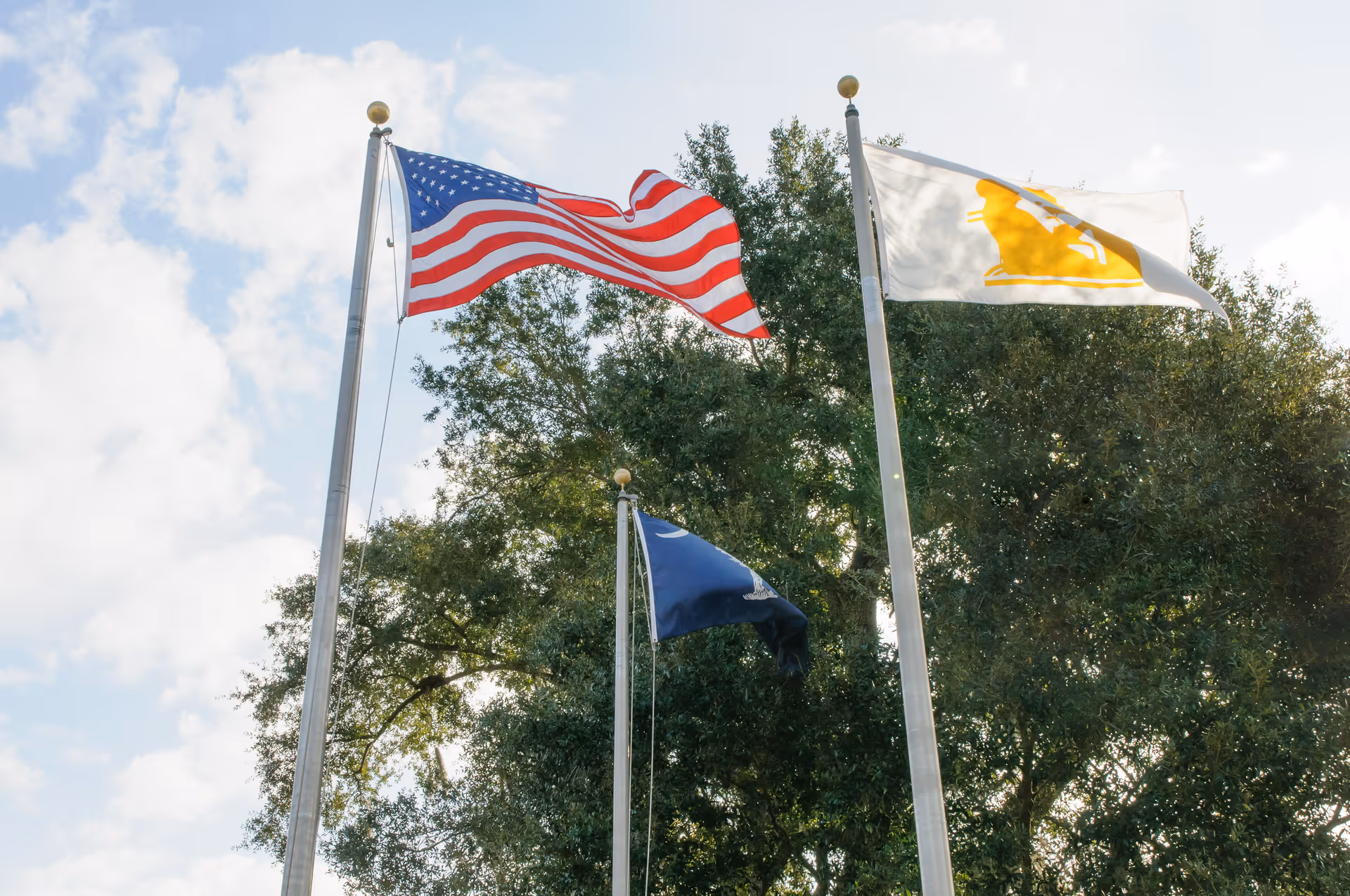 Three flags—an American flag, a dark blue flag, and a white flag with a yellow logo—flying on flagpoles in front of trees under a partly cloudy sky.