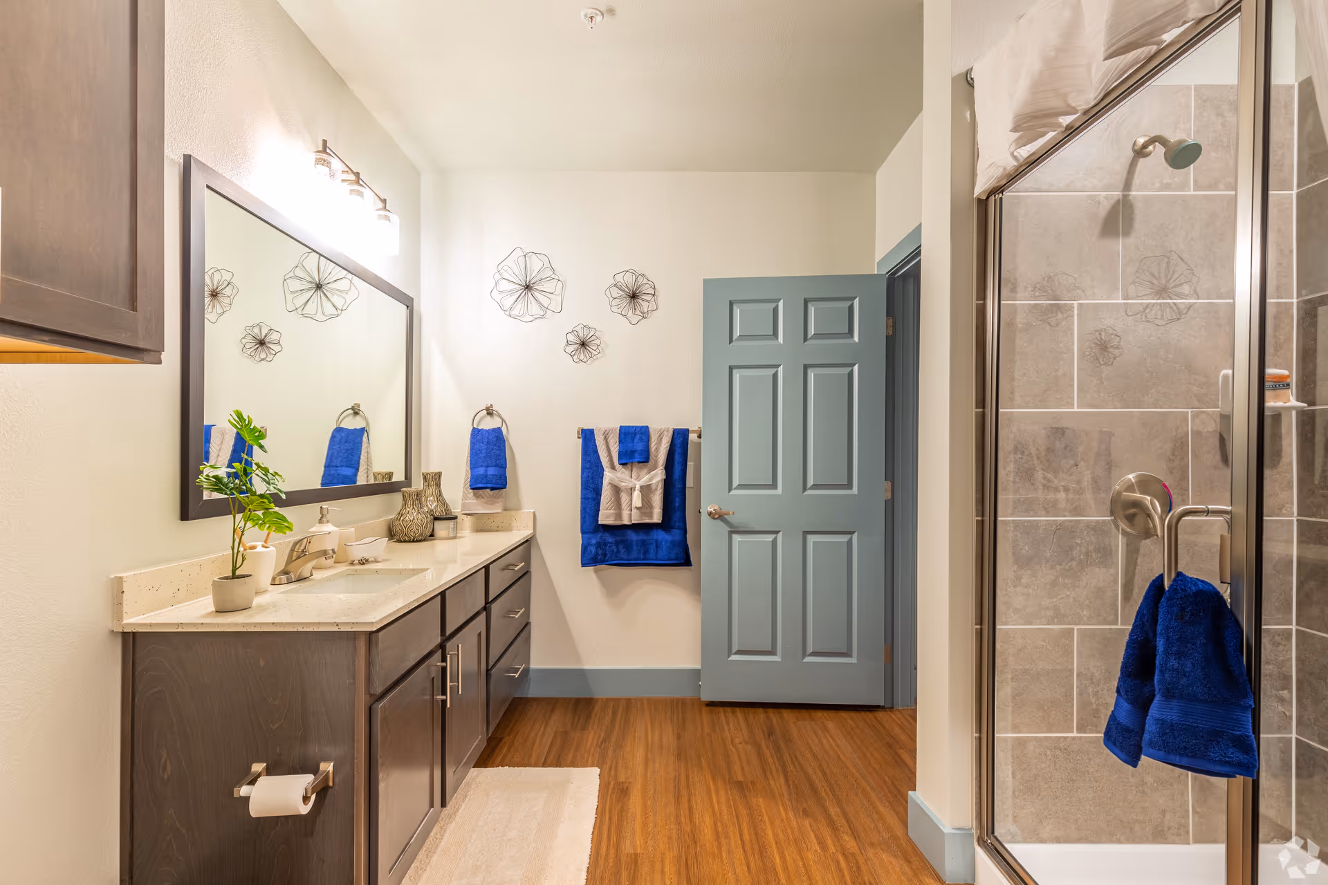 Well-lit bathroom with a double-sink vanity and large mirror, a walk-in shower, and blue towels.