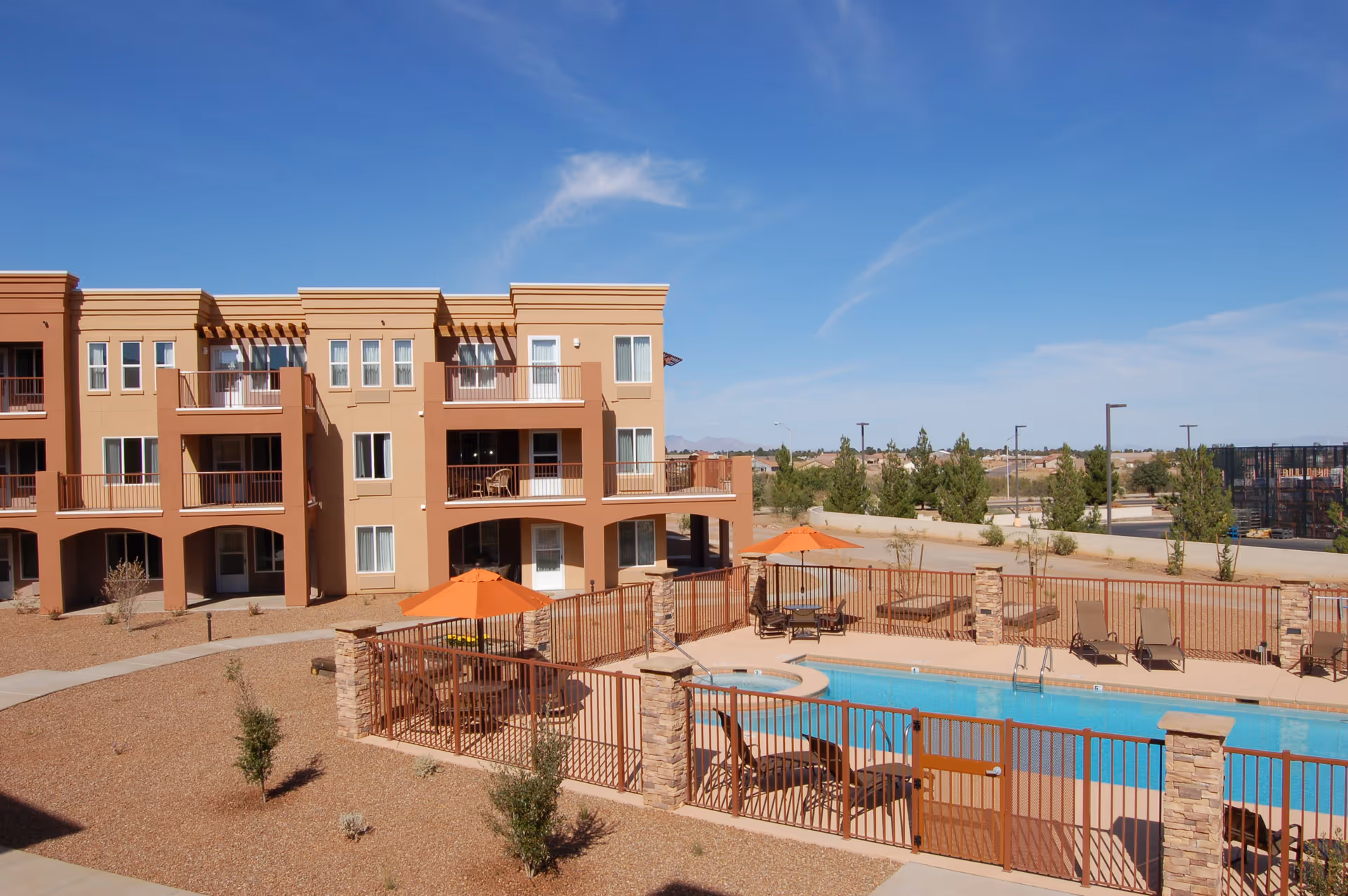 Beige three-story residential building with balconies overlooking a fenced outdoor pool and patio area under a clear blue sky.