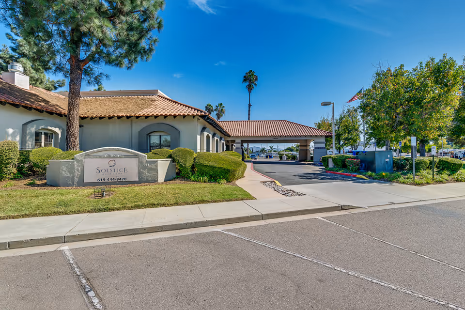 Exterior view of Solstice Senior Living at El Cajon facility showing a single-story building with a tiled roof, manicured bushes, a large tree, and a sign with the facility name and phone number. The entrance driveway is visible with a covered drop-off area, and there are trees and a flagpole with an American flag in the background under a clear blue sky.