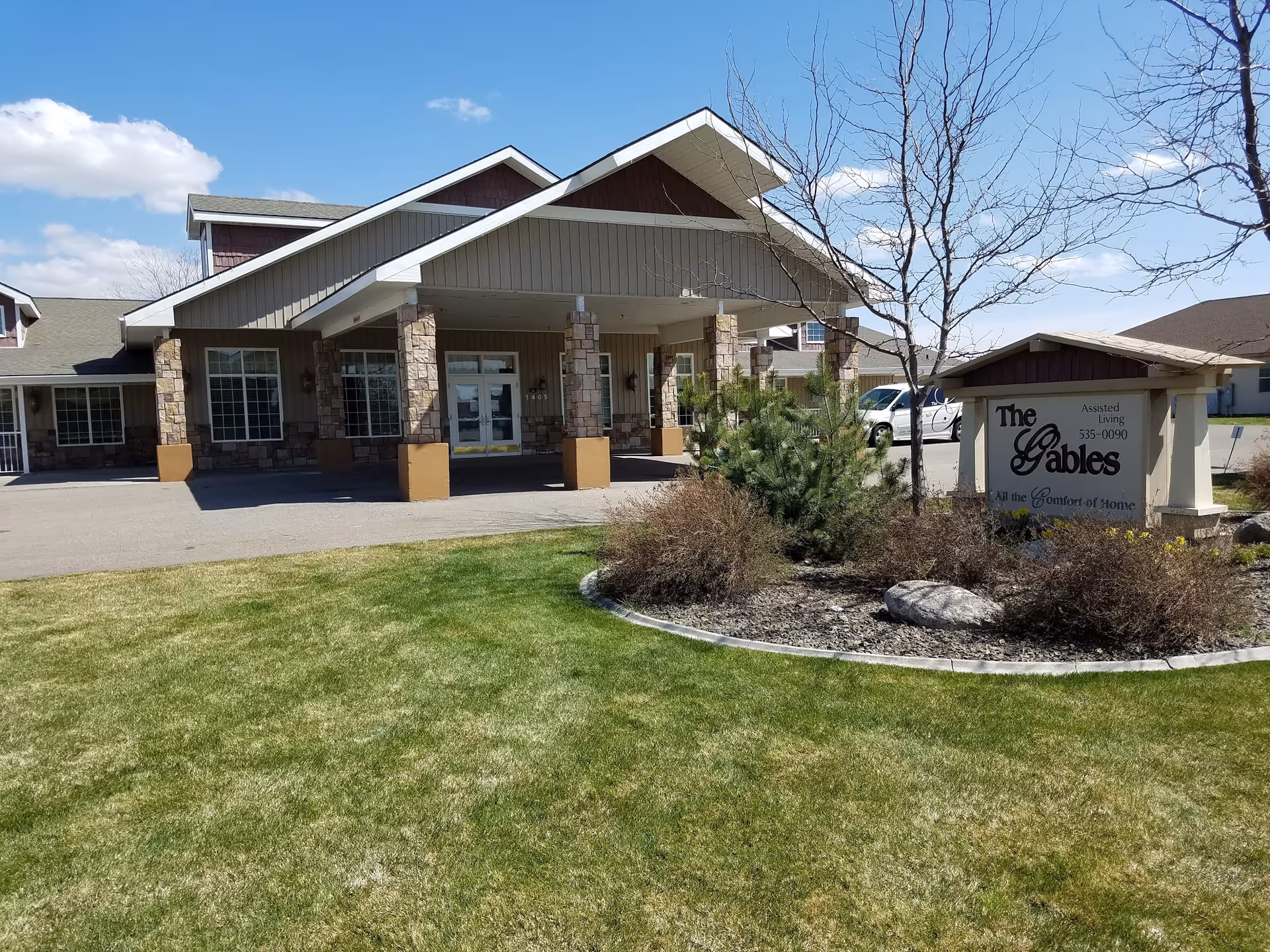 Front entrance of The Gables assisted living building with a covered porte-cochere, lawn, and a sign in the foreground.