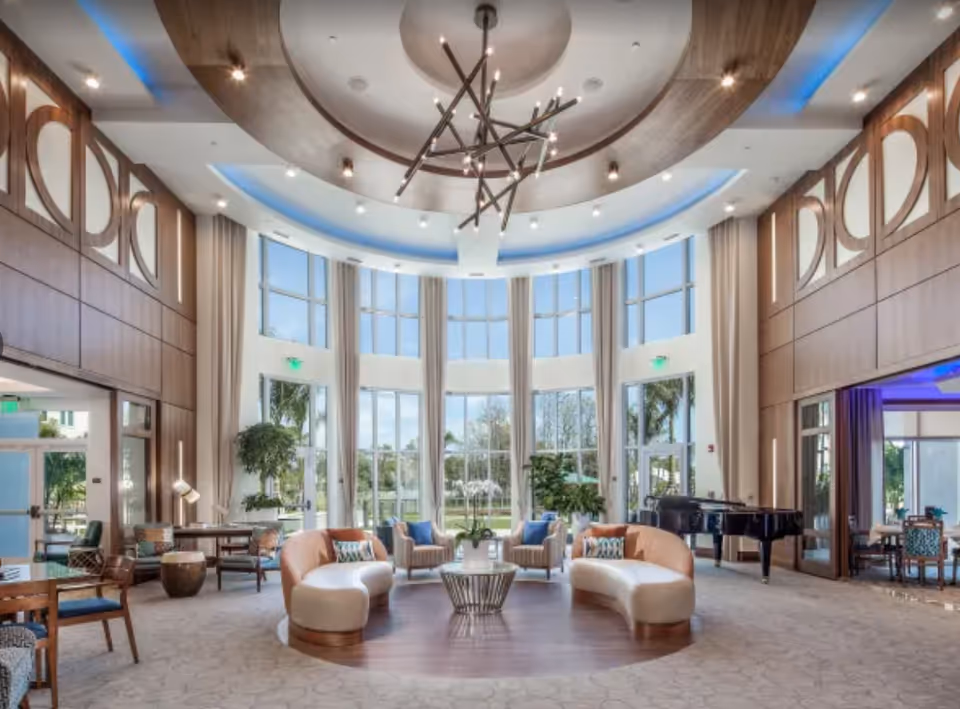 Spacious and elegant senior living facility common area with large floor-to-ceiling windows letting in natural light, curved beige sofas and armchairs arranged around a central coffee table, modern chandelier hanging from a circular ceiling design, wooden panel walls with decorative cutouts, and a black grand piano in the corner.