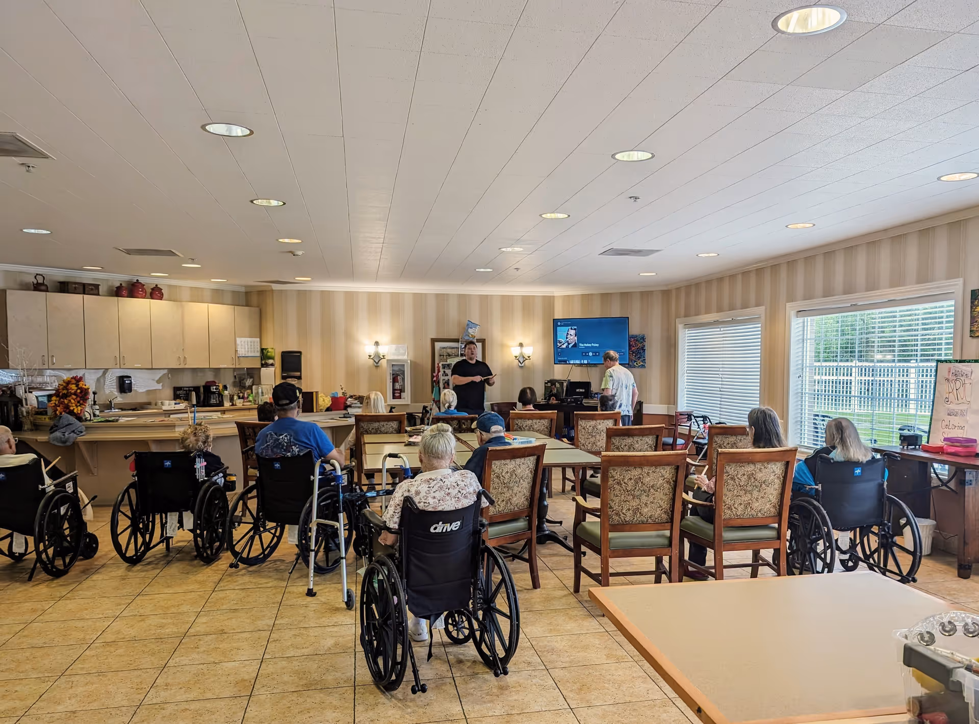 A group of elderly individuals, many in wheelchairs, seated in a common room facing a person standing and speaking near a TV mounted on the wall. The room has beige tiled floors, striped wallpaper, large windows with blinds, and a kitchenette area with cabinets and appliances on the left side.
