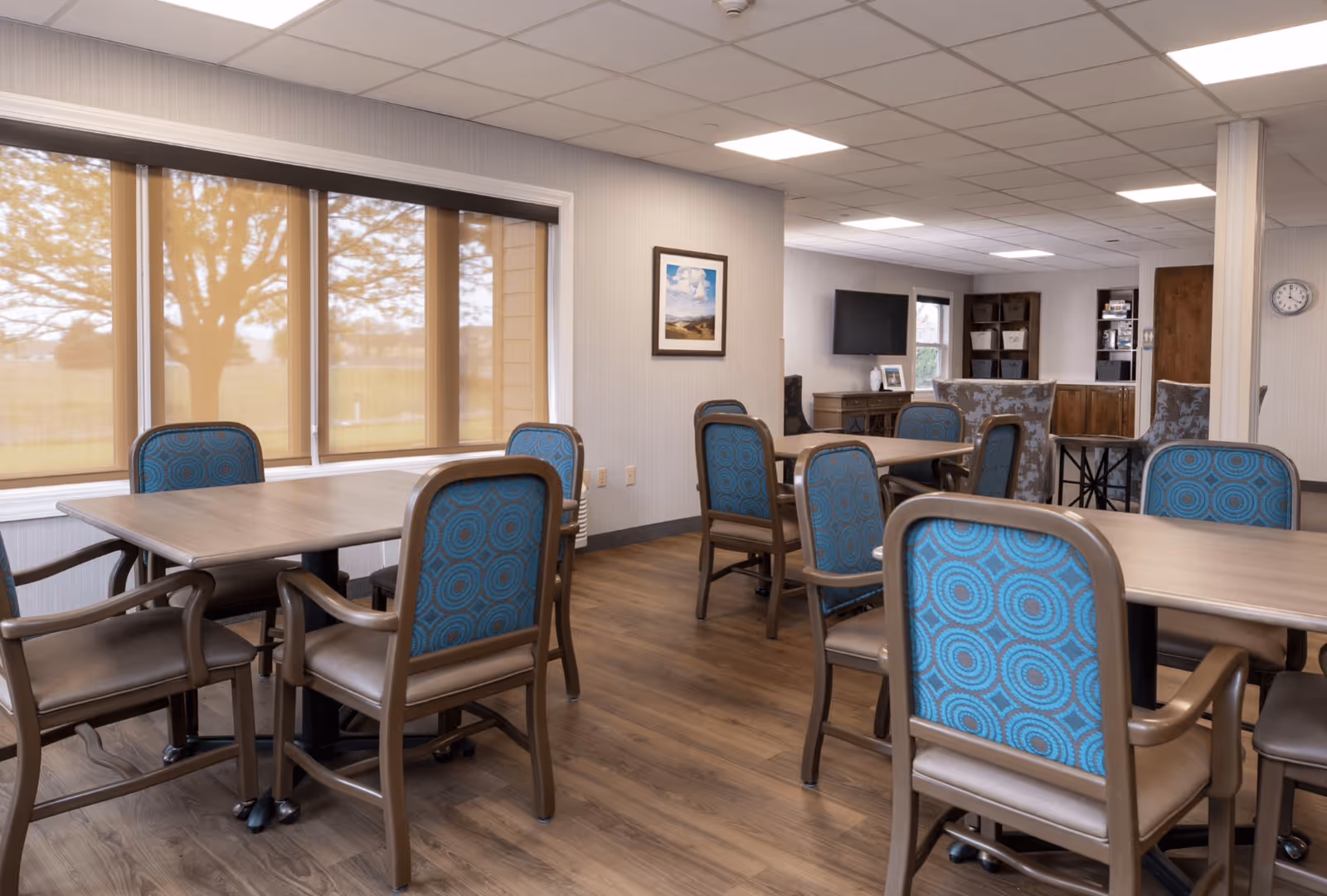 A well-lit common area with multiple wooden tables and chairs featuring blue patterned upholstery. Large windows with beige blinds allow natural light to enter, and a wall-mounted TV is visible in the background along with shelves and a clock on the wall.