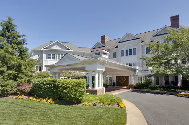 Front exterior view of a large, white senior living facility building with multiple windows, a covered entrance supported by white columns, surrounded by green trees, bushes, and a well-maintained lawn under a clear blue sky.