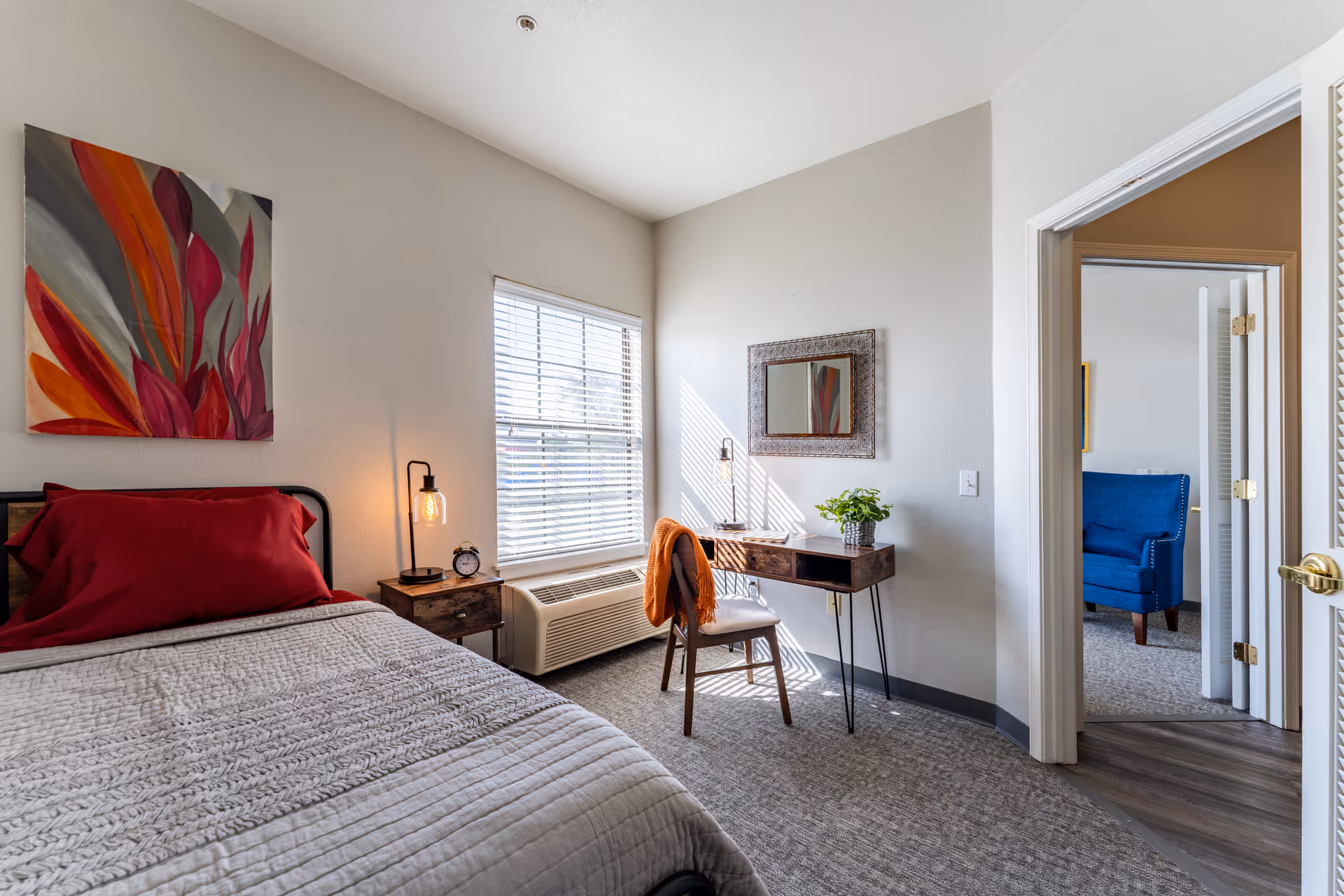 A bright bedroom with a bed featuring a red pillow and gray quilt, a wooden nightstand with a lamp and alarm clock, a window with blinds, a small wooden desk with a chair draped with an orange blanket, a decorative mirror on the wall, and an open door leading to another room with a blue armchair.