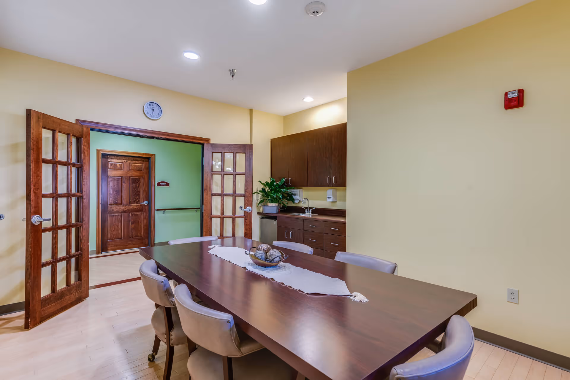 A meeting or dining room with a long wooden table surrounded by six beige cushioned chairs. The room has light yellow walls, wooden double doors with glass panes, a wall clock above the doors, and a small kitchenette area with dark wood cabinets, a sink, and a plant on the counter.