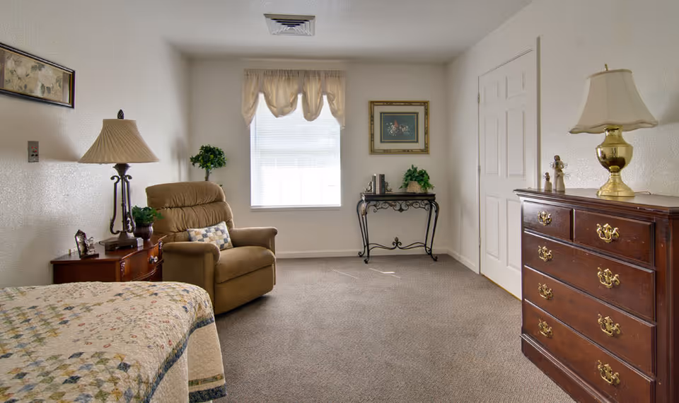 A cozy bedroom in a senior living facility featuring a bed with a patterned quilt, a comfortable recliner chair with a decorative pillow, a wooden nightstand with a lamp and small plants, a window with sheer curtains, a framed picture on the wall, a decorative table with books and a plant, and a wooden dresser with a lamp and figurines.