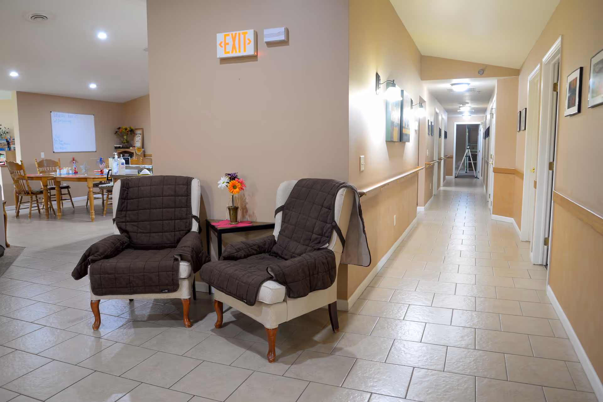 Two upholstered armchairs with dark covers and a small side table sit in a tiled hallway/common area with a dining table visible in the background and an EXIT sign overhead.