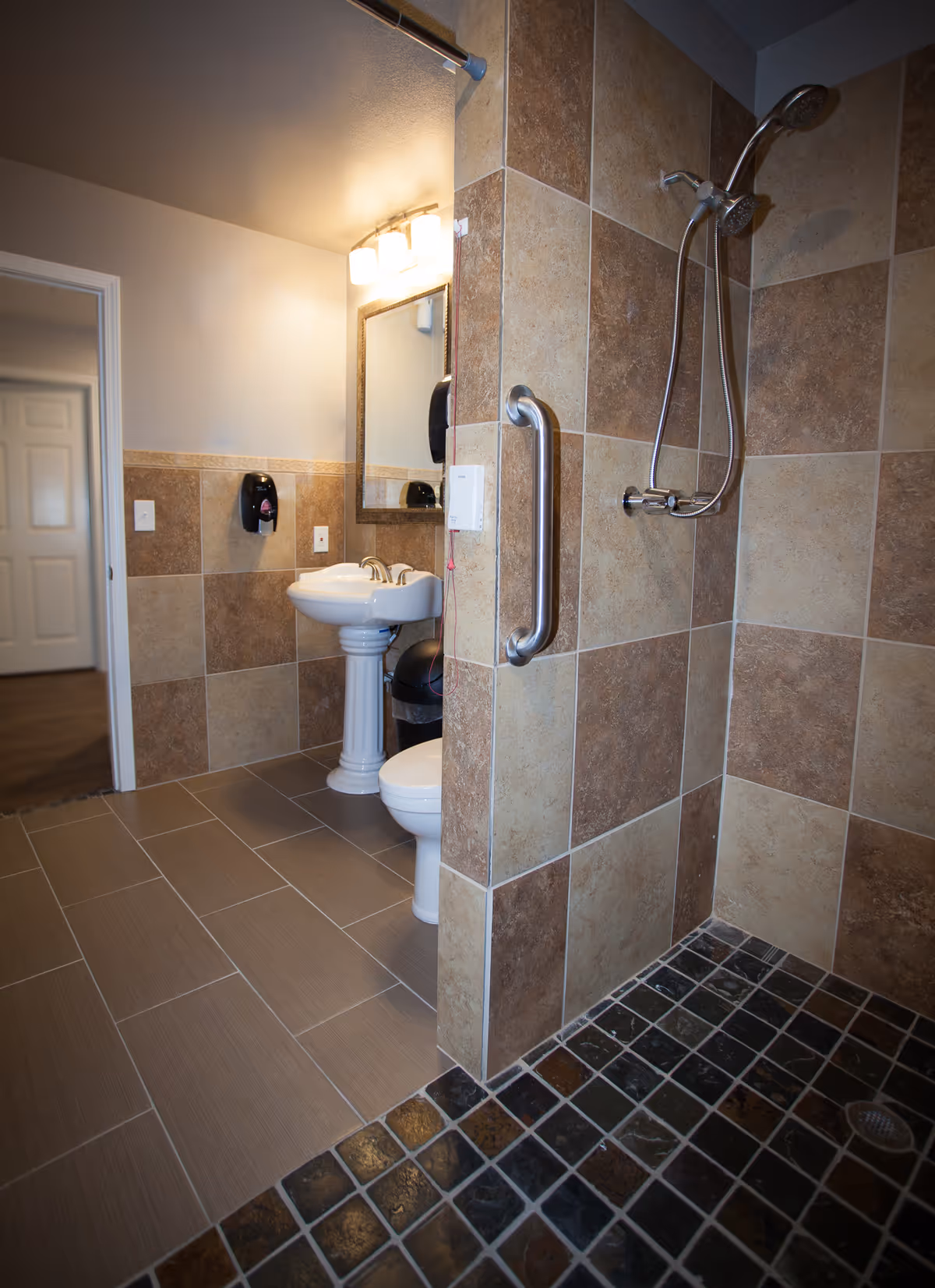 A bathroom with beige and brown tiled walls and floors. The image shows a walk-in shower with a handheld showerhead and a grab bar. Adjacent to the shower is a white pedestal sink with a mirror above it, a soap dispenser on the wall, and a toilet partially visible. The bathroom has a door leading to another room with wooden flooring.