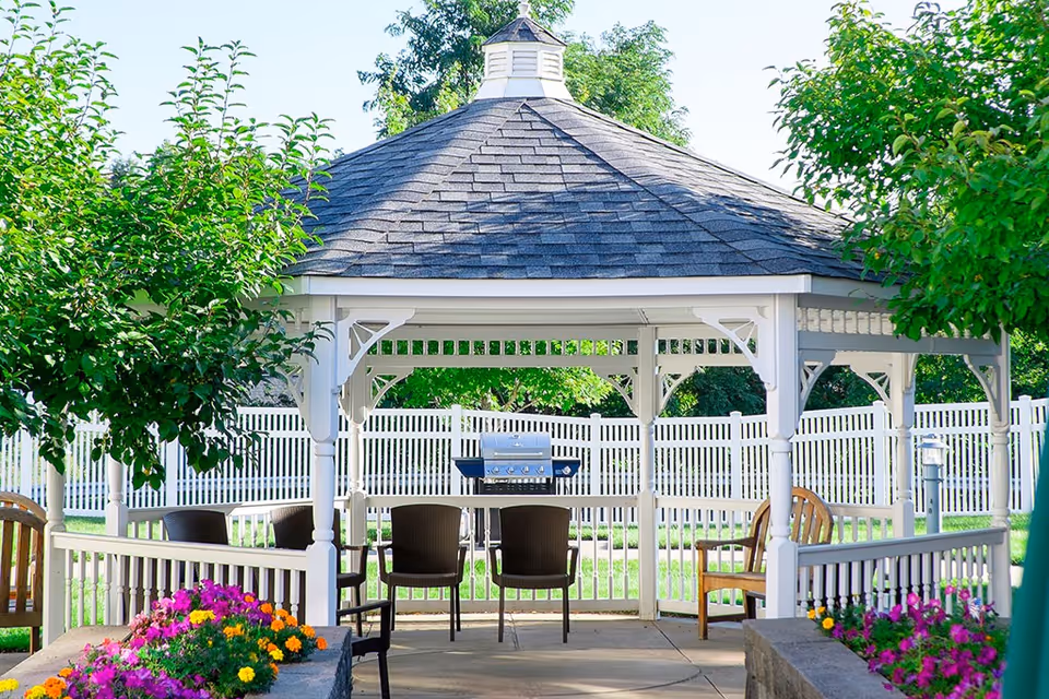 A white gazebo with a shingled roof situated in a garden area with colorful flowers and green trees. Inside the gazebo, there is a table with four chairs and a barbecue grill. The area is enclosed by a white picket fence.