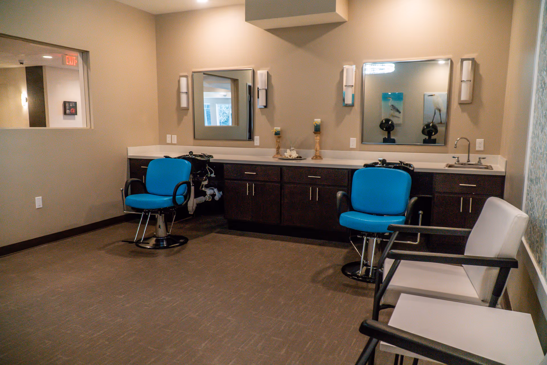 Interior room with two blue salon chairs in front of a long counter with two mirrors and a sink. There are two white chairs with black armrests along the right wall. The room has beige walls and brown flooring.