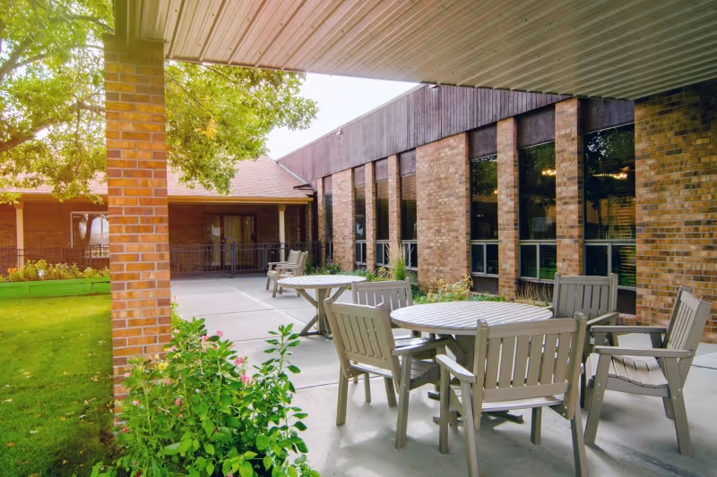 Covered outdoor patio courtyard with round tables and chairs beside a brick senior care building.