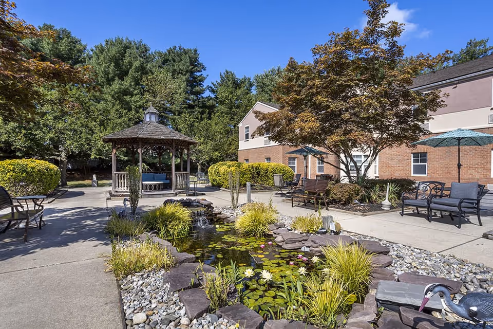 Outdoor patio area at Brookdale Evesham featuring a small pond with water lilies and a decorative heron statue, surrounded by plants and rocks. There is a wooden gazebo with seating, several benches, chairs, and umbrellas. The background shows a brick building and trees under a clear blue sky.