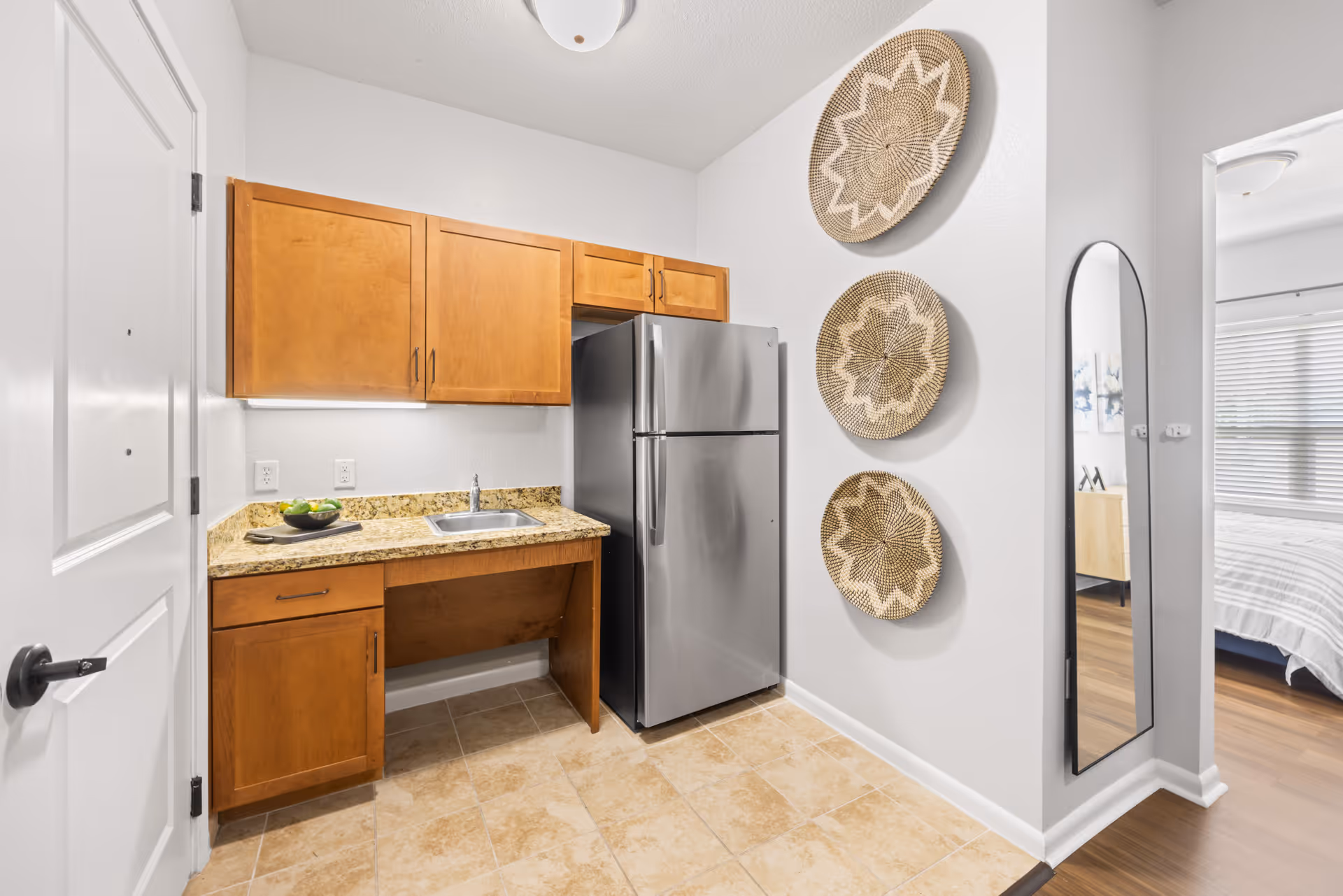 A small kitchen area with wooden cabinets, a granite countertop with a sink, and a stainless steel refrigerator. On the wall to the right, three decorative woven baskets are hung vertically above a tall, narrow mirror. The floor is tiled, and a doorway leads to a bedroom with a bed and window blinds visible.