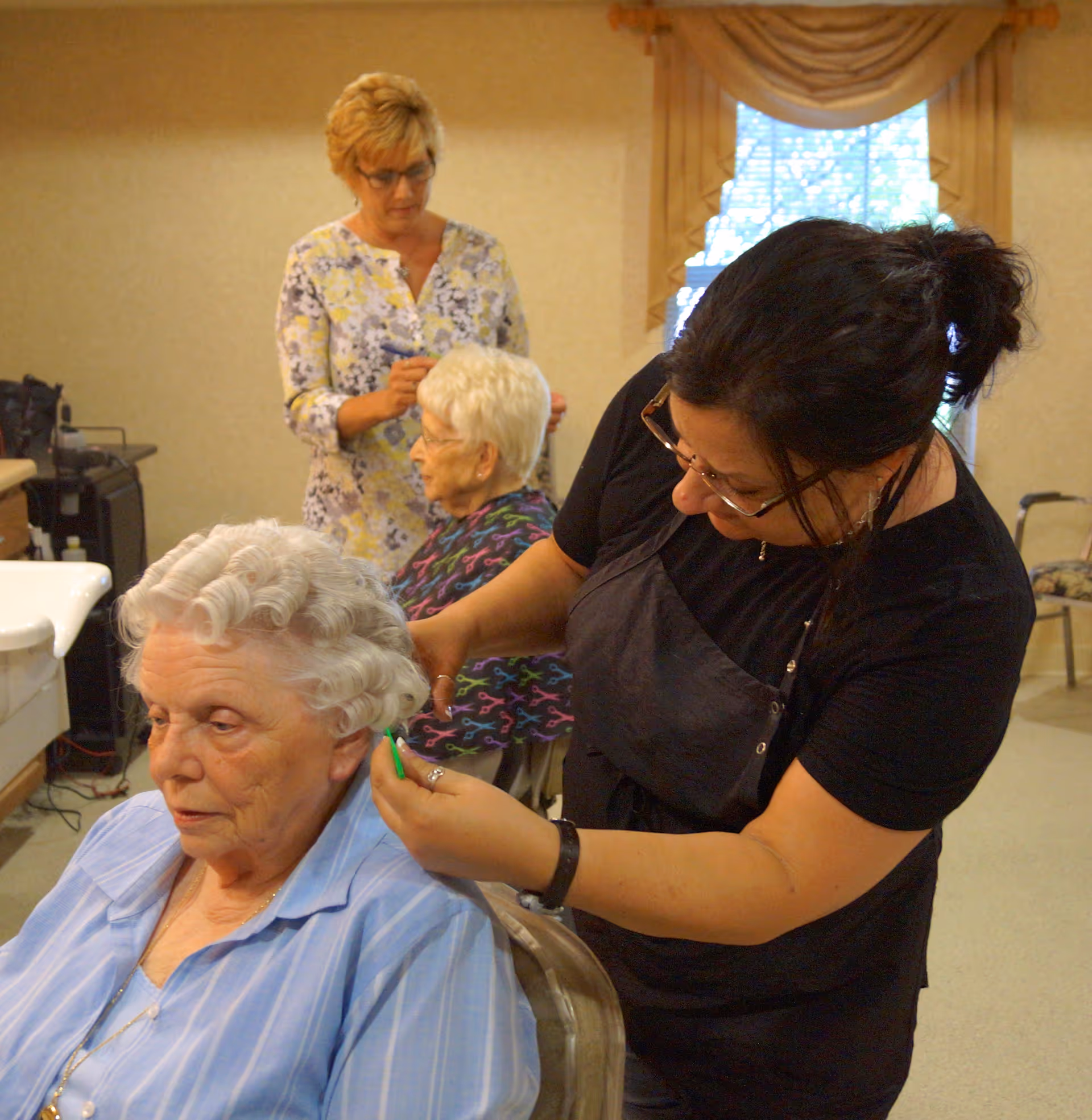 A hairstylist is curling the hair of an elderly woman seated in a chair, while another elderly woman is seated in the background with a caregiver standing behind her. The setting appears to be a cozy room with beige walls and a window with drapes.